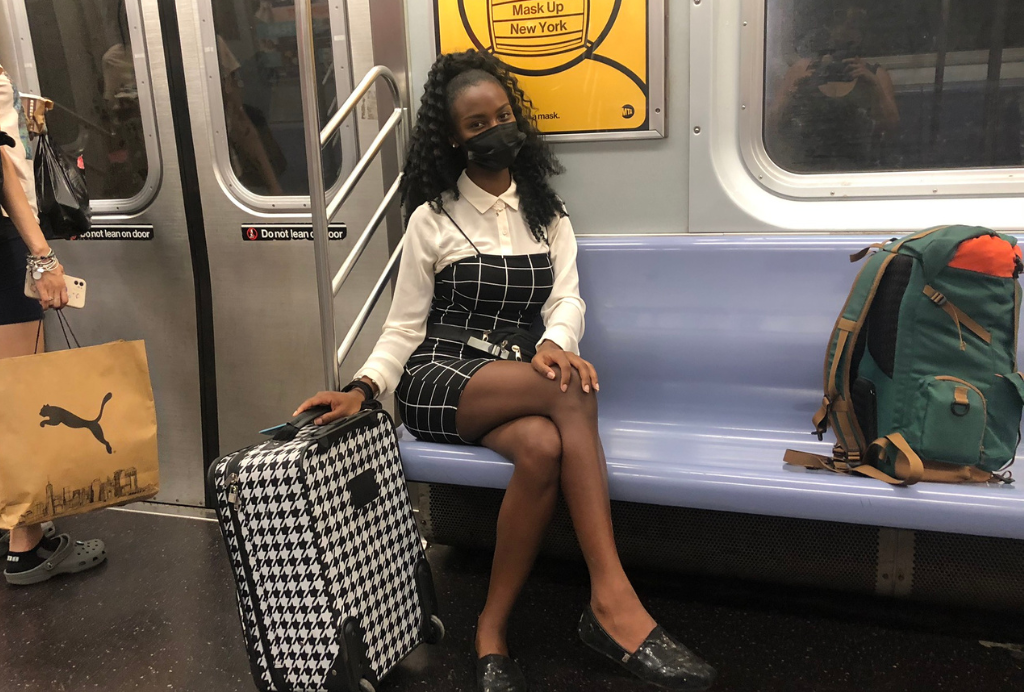 A black woman on a subway in new York with a carry-on rolling suitcase and a full back pack on the seat next to her. 